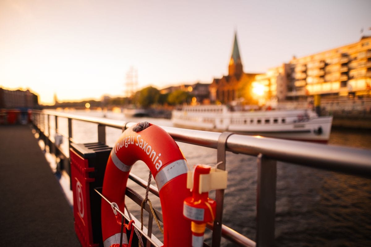 Ein Rettungsring mit der Aufschrift 'Thurgau Saxonia' an einem Steg, im Hintergrund sind Boote und die Stadtansicht bei Sonnenuntergang sichtbar.
