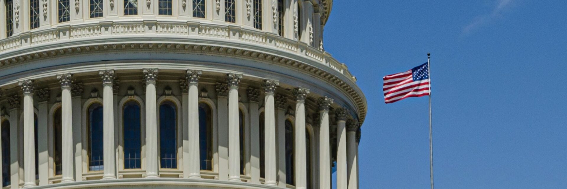 Detailansicht des Kapitols in Washington D.C. mit Säulen und einer wehenden amerikanischen Flagge vor klarem Himmel.