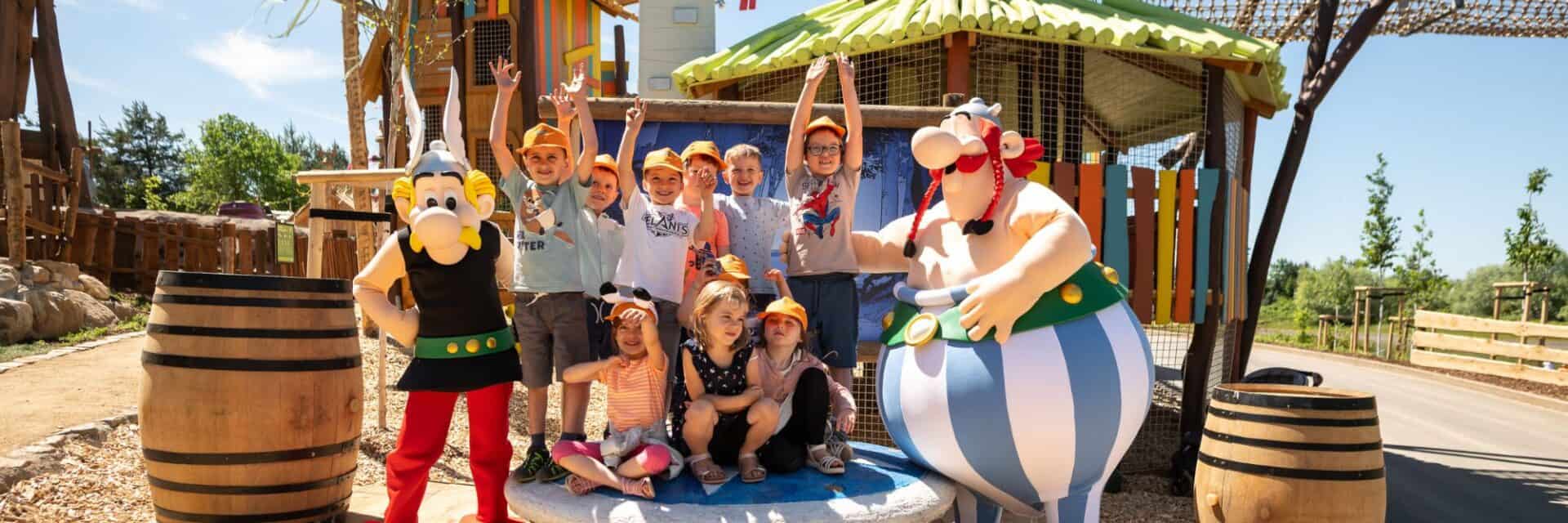 Eine Gruppe von Kindern in T-Shirts und Hüten posiert fröhlich mit zwei Comicfiguren vor einem bunten Spielplatz im Freien.