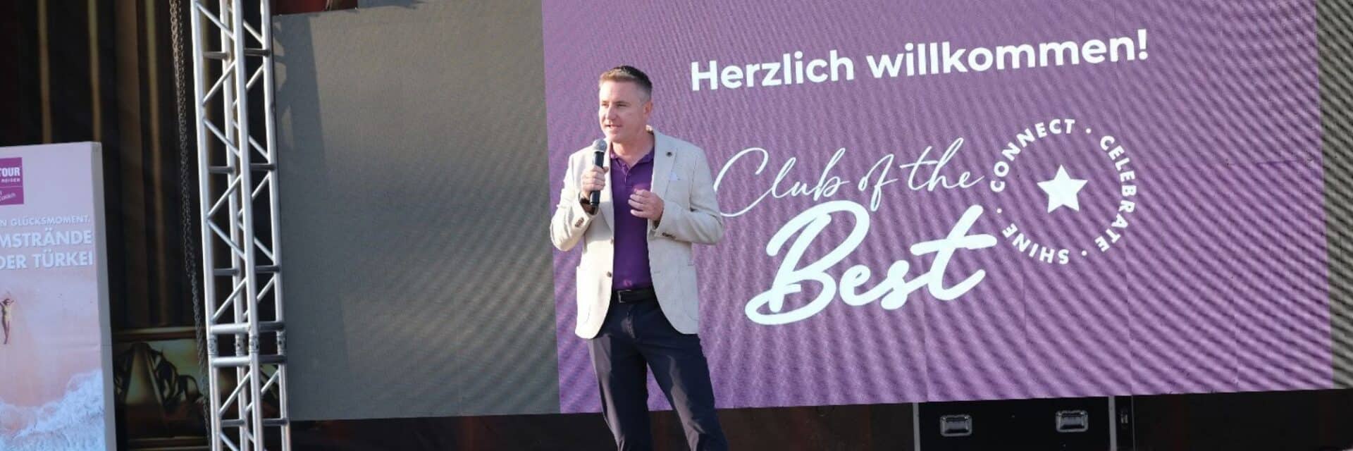 Ein Mann in heller Jacke und dunkler Hose steht auf einer Bühne und spricht in ein Mikrofon, im Hintergrund ein großes Banner mit Willkommensbotschaft.