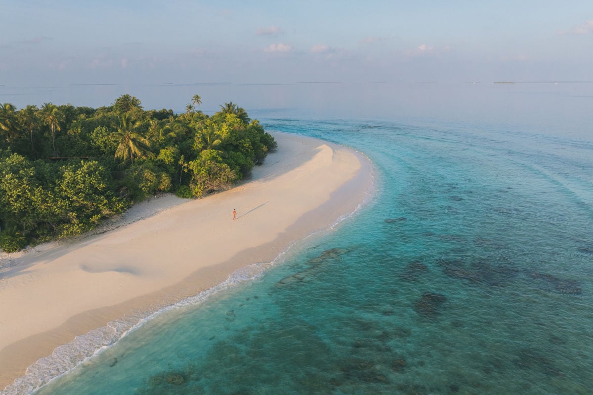 Blick aus der Luft auf eine tropische, grün bewachsene Insel im Abendlicht. Eine Frau läuft auf dem Sandstrand entlang.