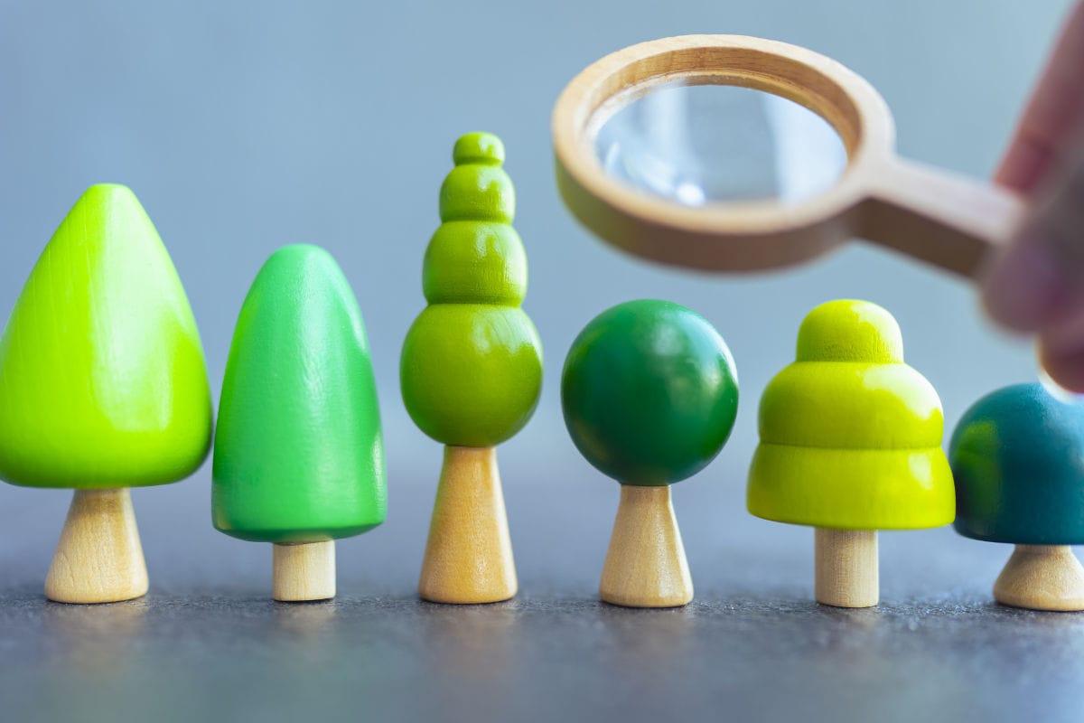 Mehrere kleine, grün bemalte Holzfiguren in Form stilisierter Bäume stehen nebeneinander auf einer Oberfläche. Rechts hält eine Hand eine Lupe über die Figuren, als würde sie diese genauer betrachten oder untersuchen.