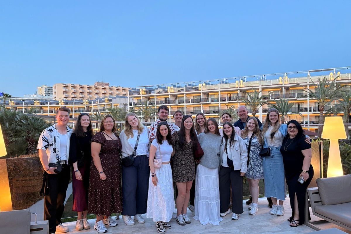 Gruppe von Reisebüro-Expedienten posiert vor einem modernen Hotel mit Palmen im Hintergrund während eines Branchenevents.