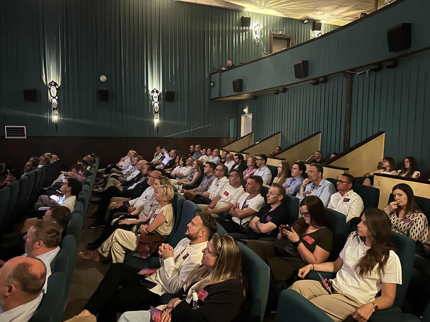 Eine Gruppe von Menschen sitzt in einem Kino, aufmerksam auf die Leinwand gerichtet. Die Zuschauer sind in Reihen angeordnet, einige mit Notizen.
