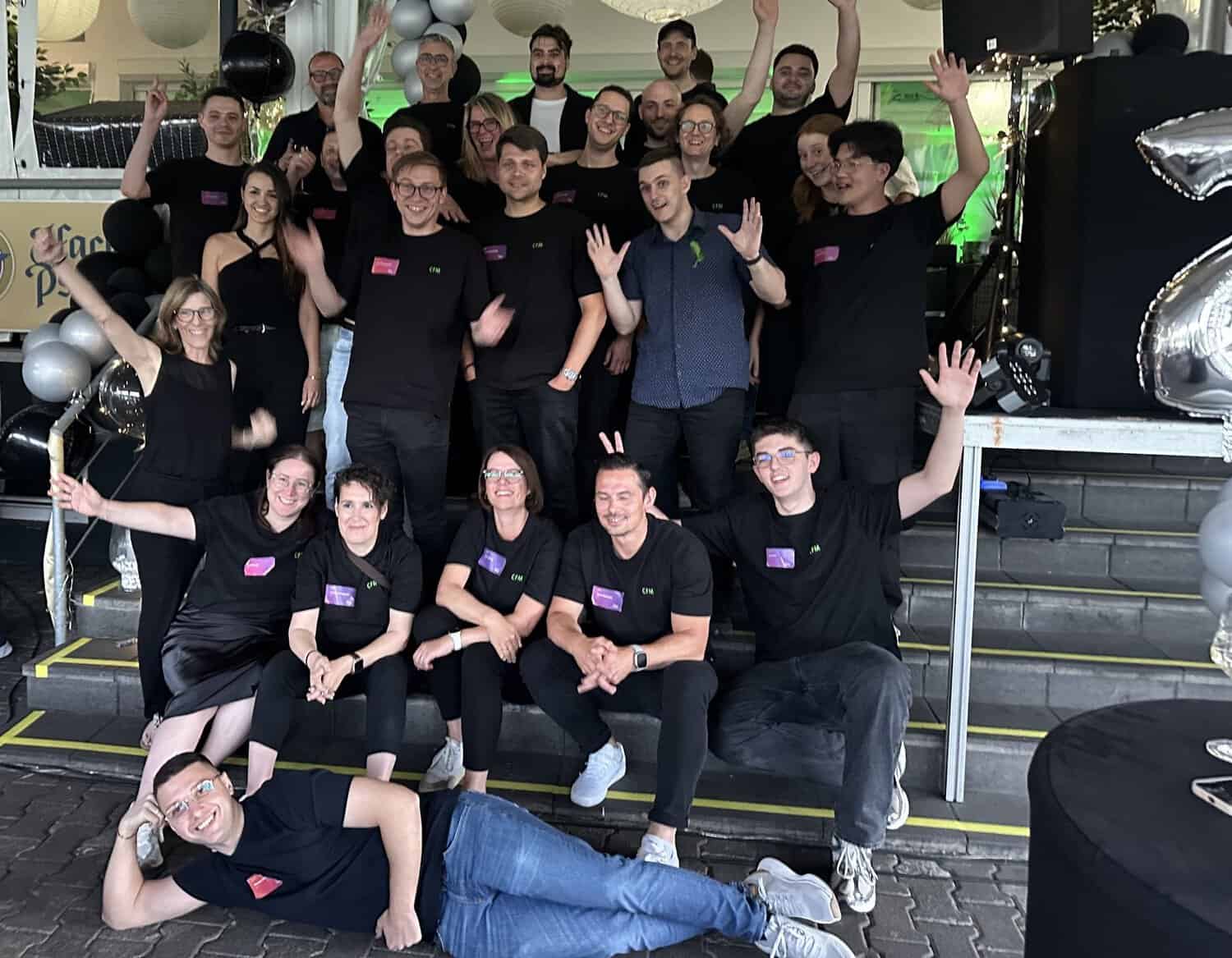 Eine Gruppe von 20 Personen in schwarzen T-Shirts posiert fröhlich vor einer Veranstaltung, umgeben von Luftballons und Dekoration.
