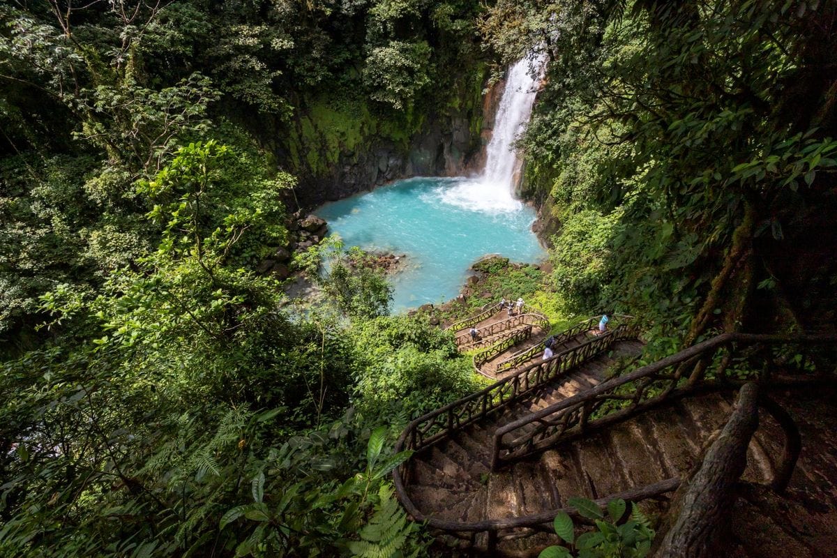 Eine malerische Landschaft mit einem Wasserfall, umgeben von üppigem Grün und einem klaren, blauen Wasserbecken, sowie geschwungenen Holztreppen.