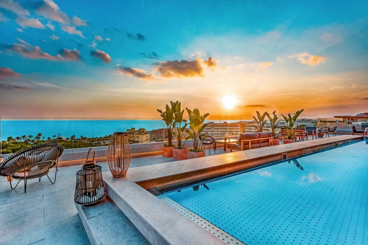 Ein eleganter Infinity-Pool auf einer Dachterrasse mit Blick auf das Meer und einen Sonnenuntergang, umgeben von modernen Möbeln und Pflanzen.