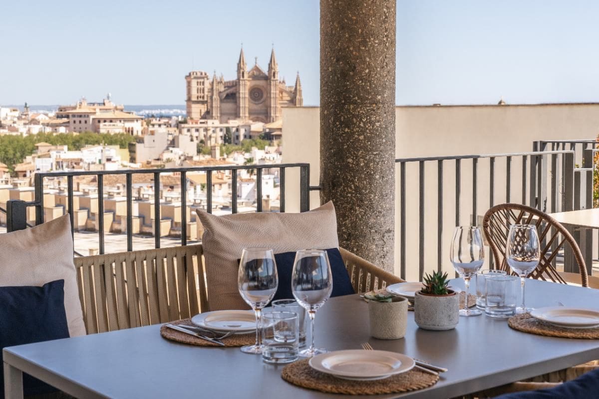 Ein eleganter Tisch auf einer Terrasse mit Blick auf die Stadt Palma de Mallorca, dekoriert mit Gläsern, Tellern und Pflanzen, im Hintergrund eine Kathedrale.
