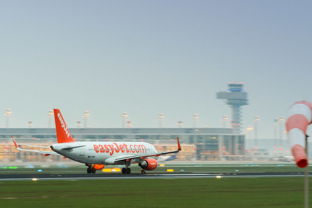 Ein EasyJet-Flugzeug rollt auf einer Landebahn, im Hintergrund sind das Terminal und der Kontrollturm eines Flughafens sichtbar.