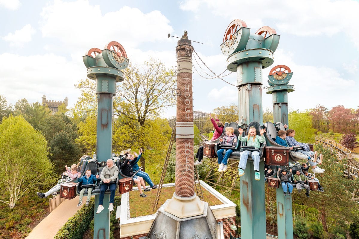 Mehrere Personen sitzen in einer Freizeitpark-Attraktion mit rotierenden Sitzreihen, die an hohen, türkisfarbenen Säulen befestigt sind. Die Fahrgäste lachen, winken oder halten sich fest, während die Sitze in der Luft schweben. In der Mitte steht ein hoher, schornsteinartiger Turm, umgeben von Bäumen und weiteren Fahrgeschäften im Hintergrund.