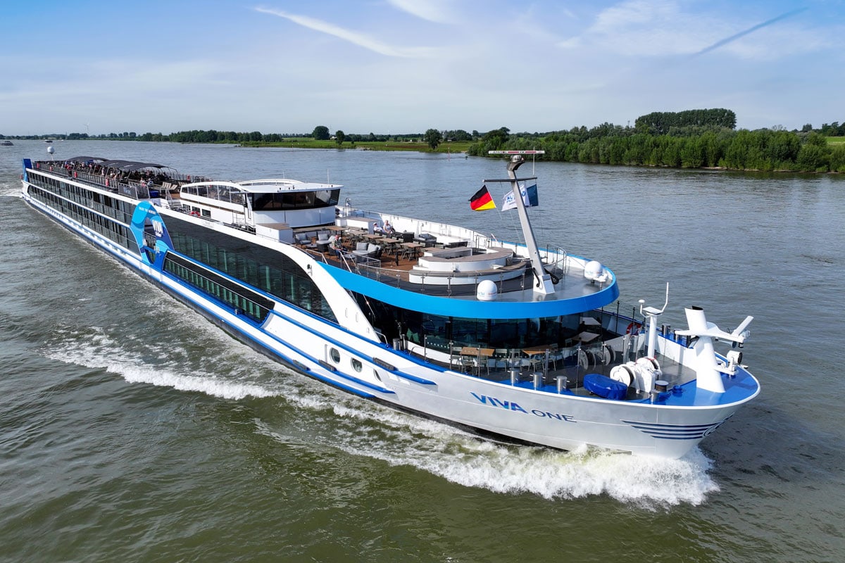 Ein modernes Flusskreuzfahrtschiff mit blauer und weißer Farbgestaltung fährt auf einem ruhigen Fluss, umgeben von grüner Ufervegetation.