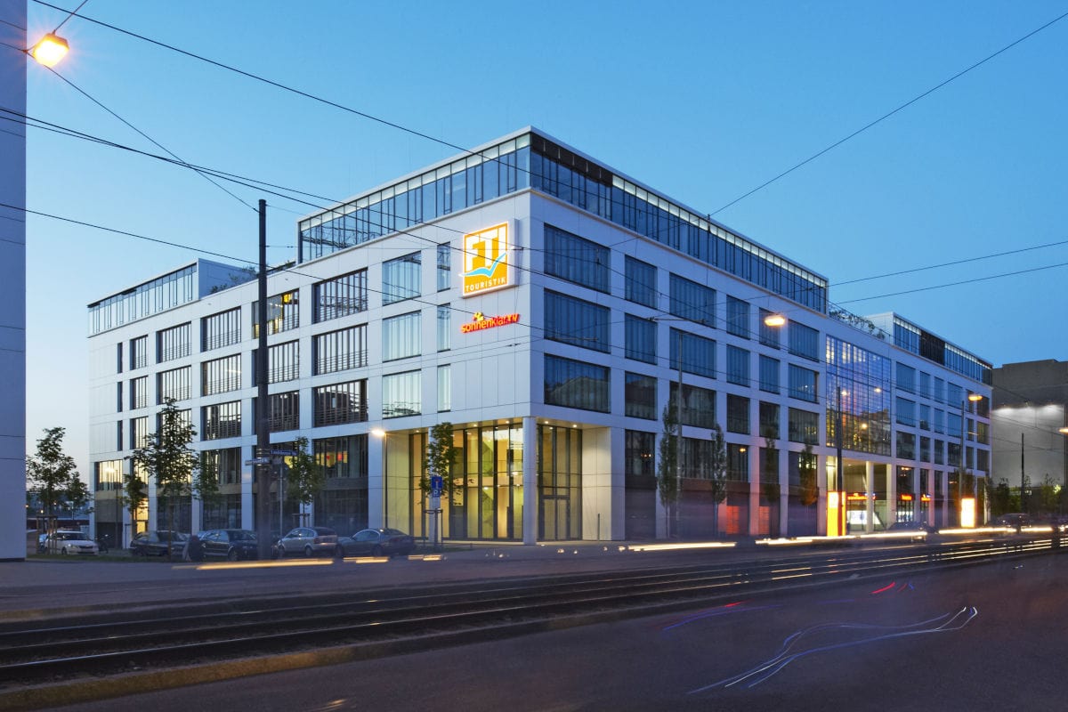 Das Bild zeigt die moderne Firmenzentrale von FTI Touristik in München bei Abenddämmerung, mit großen Glasfronten und dem beleuchteten FTI-Logo an der Fassade. Im Vordergrund verlaufen Straßenbahngleise und einige Autos sind geparkt.