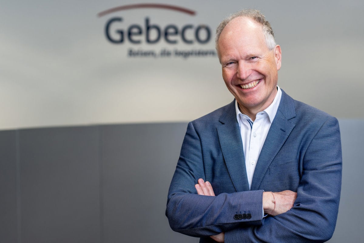 Ein lächelnder Mann in einem blauen Anzug steht mit verschränkten Armen vor einer grauen Wand mit dem Logo von Gebeco.