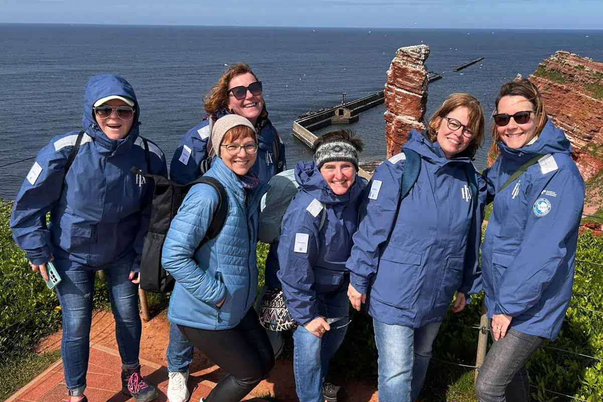 Sechs Reisebüro-Expedienten posieren vor einer Küstenlandschaft mit Felsen und Meer im Hintergrund.