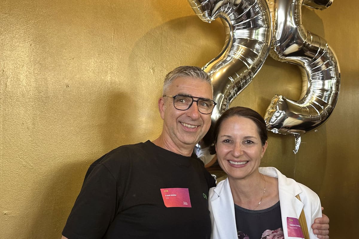 Zwei Personen posieren vor einer goldenen Wand, mit silbernen Luftballons in Form der Zahl 25 im Hintergrund. Beide lächeln freundlich.