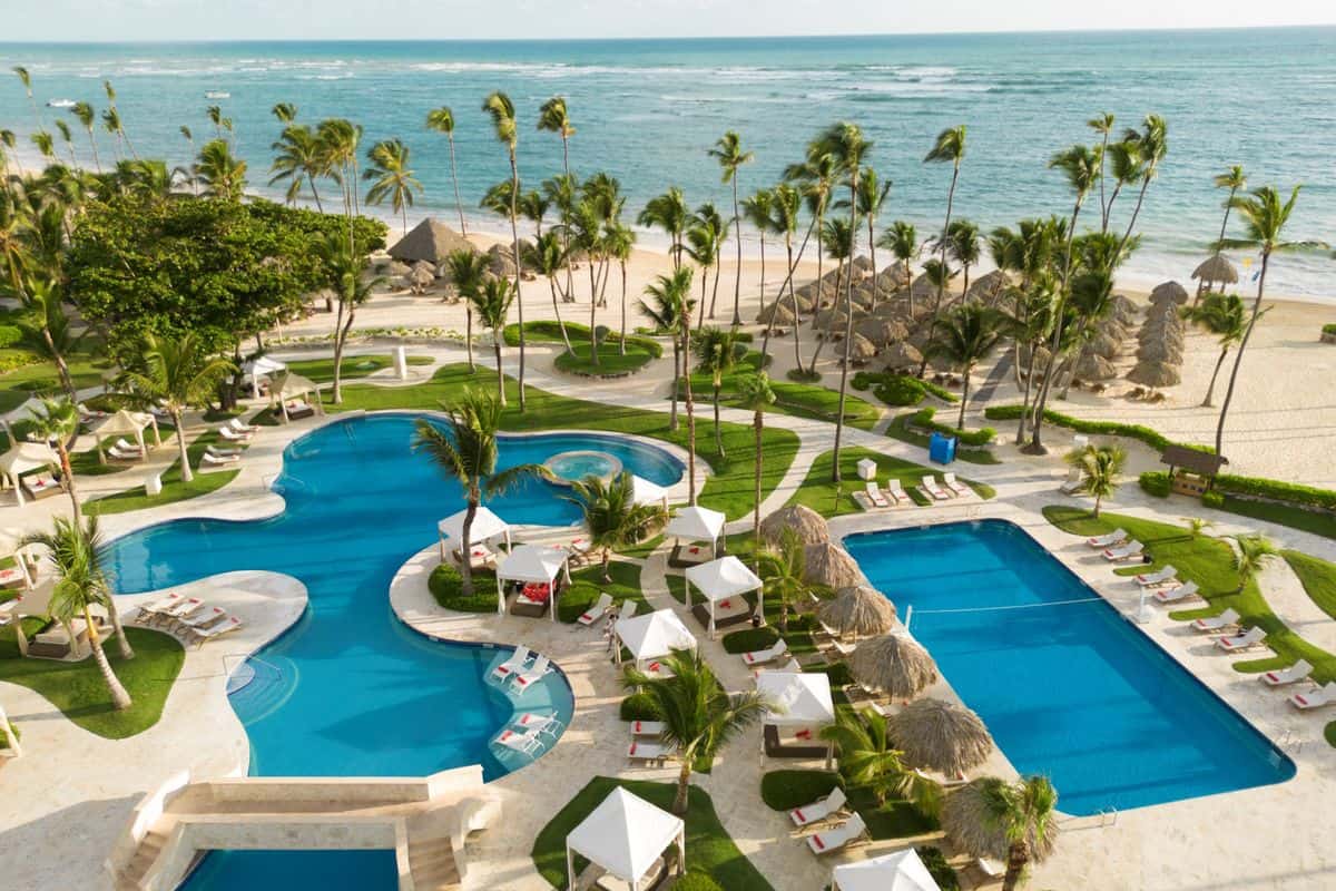 Luftaufnahme eines luxuriösen Resorts mit zwei Pools, Liegen und Palmen, umgeben von einem Strand und dem Meer im Hintergrund.