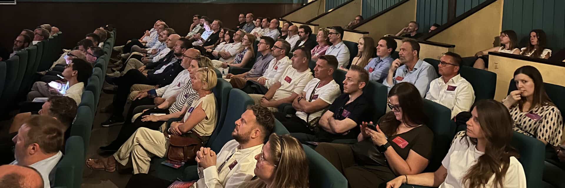 Eine große Gruppe von Menschen sitzt in einem Theater, aufmerksam auf die Bühne gerichtet, in einem modernen Veranstaltungsraum.