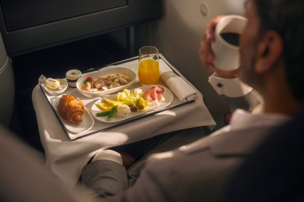 Ein Mann genießt in der Business Class von Lufthansa ein luxuriöses Frühstücksmenü, bestehend aus Croissant, Aufschnitt, Käse, Müsli, Butter, Konfitüre, Orangensaft und einer Tasse Kaffee auf einem Tablett.