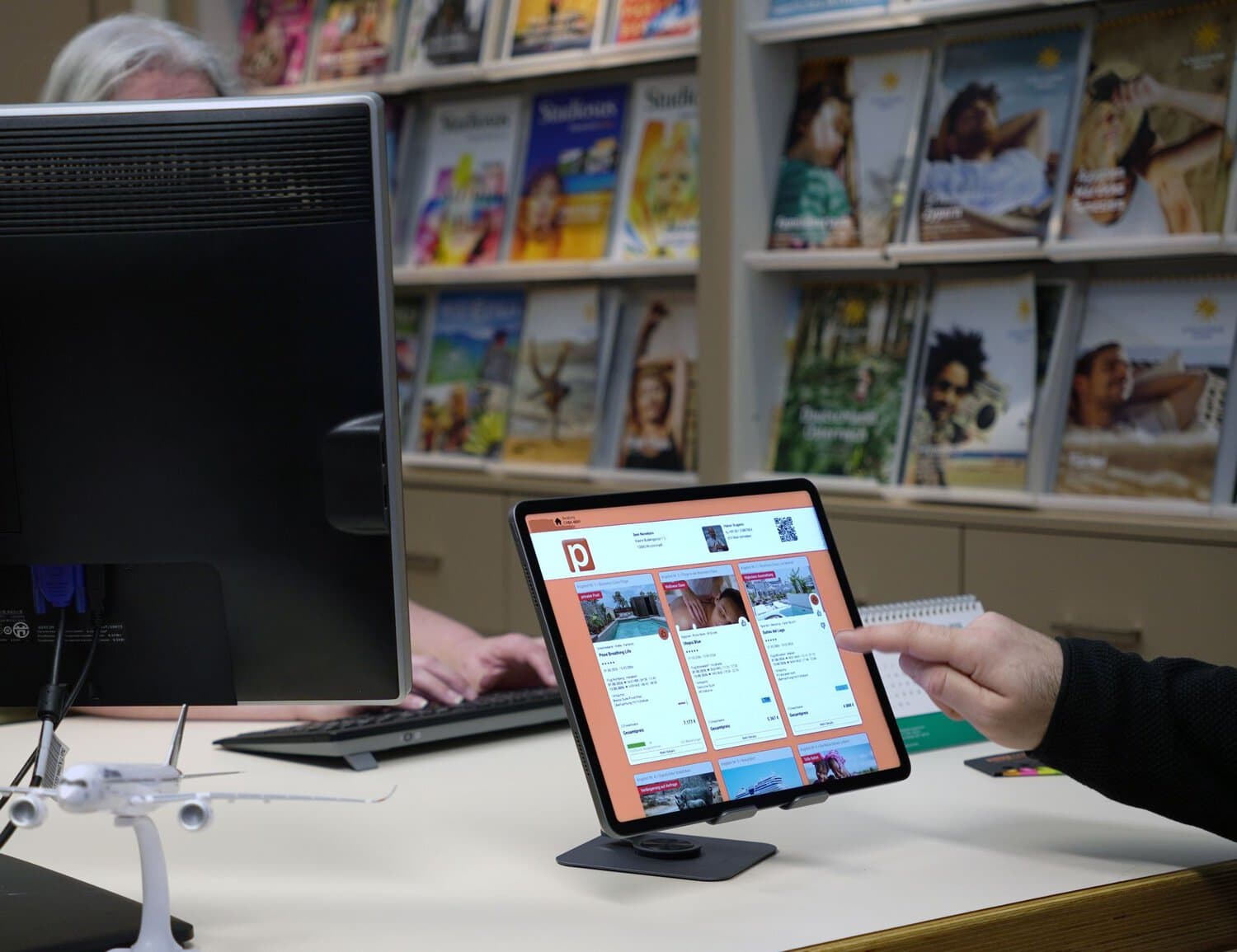 Eine Person zeigt mit dem Finger auf ein Tablet auf einem Schreibtisch. Auf dem Bildschirm ist eine Reise- und Buchungsplattform mit verschiedenen Hotel- und Urlaubsangeboten zu sehen. Im Hintergrund stehen ein Monitor und eine Tastatur, im Vordergrund befindet sich ein kleines Flugzeugmodell.