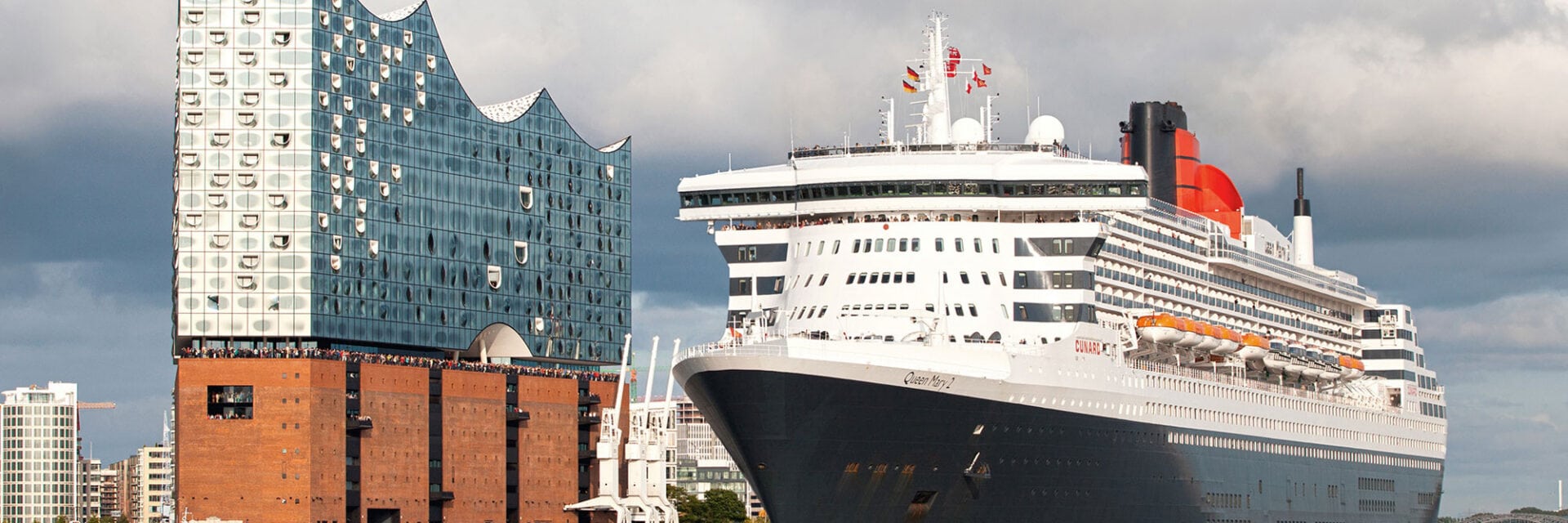 Ein Kreuzfahrtschiff liegt im Hafen vor einem modernen, gläsernen Gebäude mit wellenförmiger Fassade und einer historischen Backsteinstruktur.