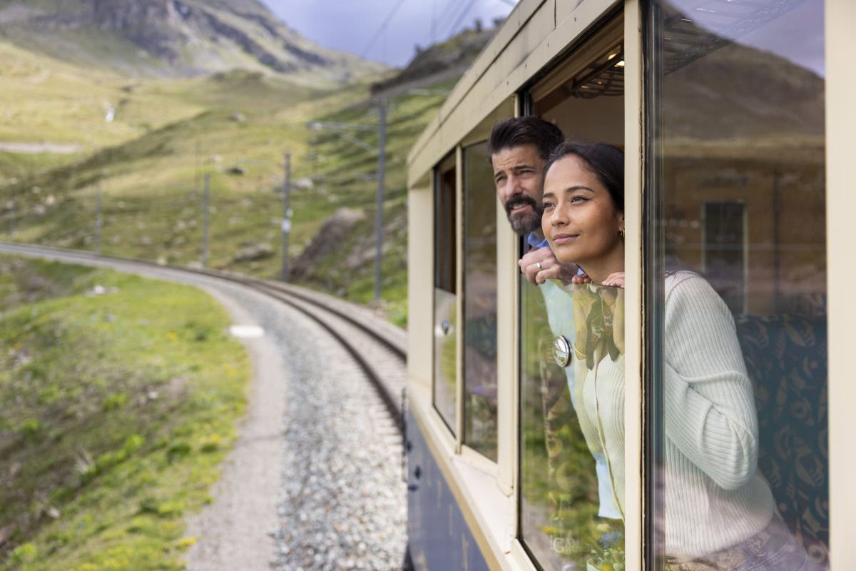 Zwei Personen lehnen am geöffneten Fenster eines fahrenden Zuges und blicken auf eine kurvenreiche Bahnstrecke durch eine grüne Berglandschaft. Im Hintergrund sind sanfte Hänge, Felsen und Strommasten entlang der Gleise zu sehen.