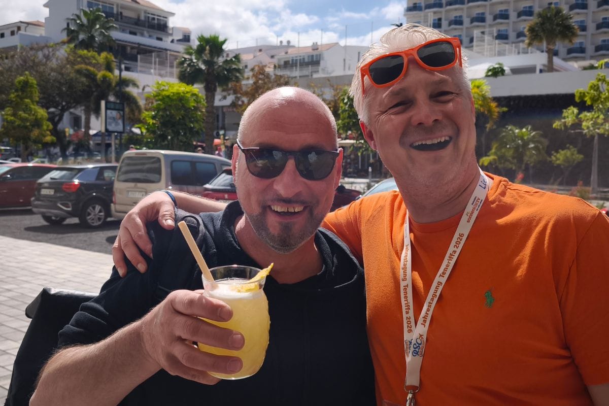 Das Foto zeigt zwei Personen, die gemeinsam einen sonnigen Tag im Urlaubsort Teneriffa genießen; einer der Männer hält einen frischen Cocktail, während der andere ein orangefarbenes Polohemd und ein Tagungsband trägt. Im Hintergrund sind Palmen, parkende Autos und moderne Hotelgebäude sichtbar.