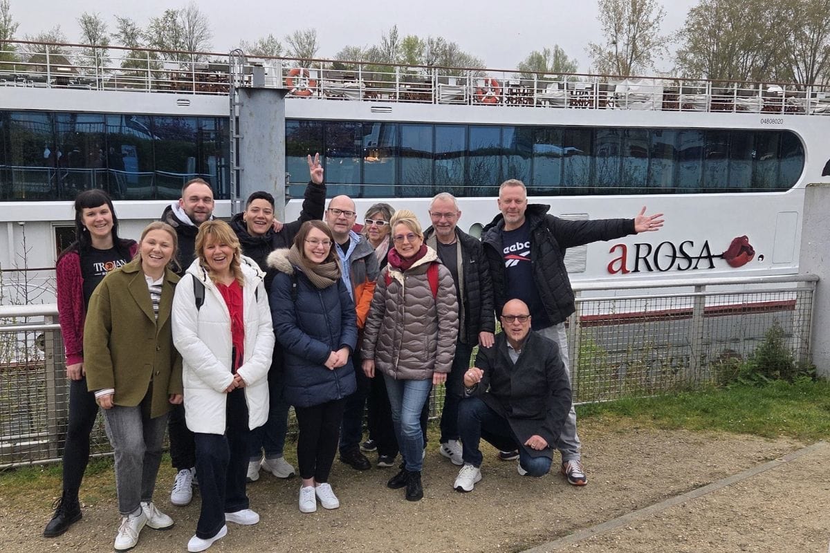 Gruppe von Reisebüro-Expedienten vor einem A-Rosa Kreuzfahrtschiff, lächelnd und in lockerer Atmosphäre.