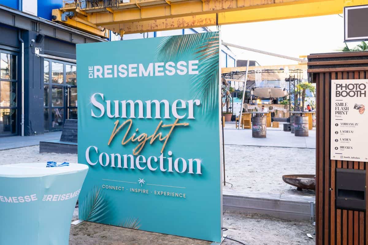 Ein dekoratives Banner mit der Aufschrift 'Summer Night Connection' vor einem Veranstaltungsort, umgeben von Sand und Sitzgelegenheiten.