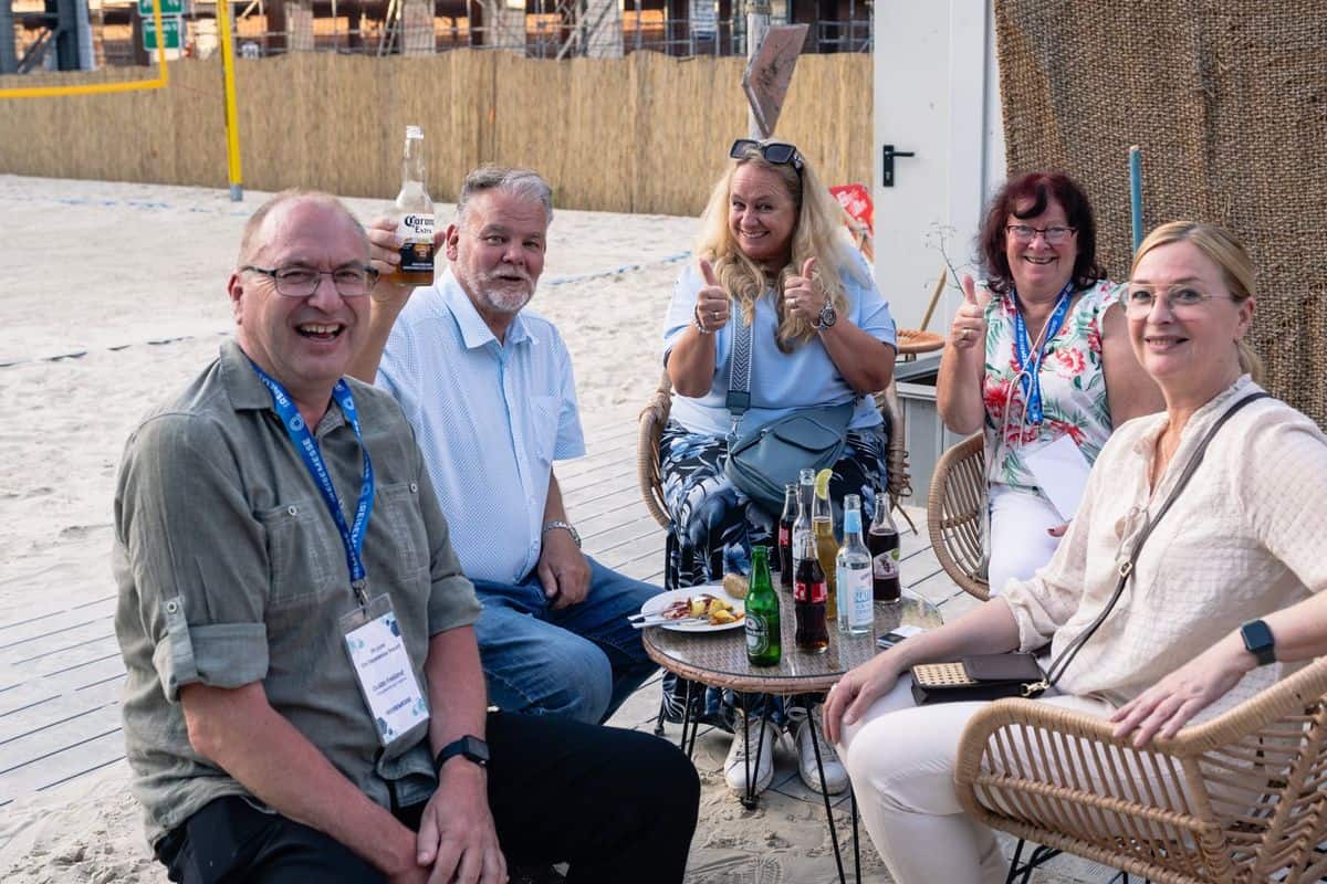 Fünf Personen sitzen entspannt an einem Tisch am Strand, umgeben von Sand und einer natürlichen Kulisse. Getränke und Snacks sind auf dem Tisch.
