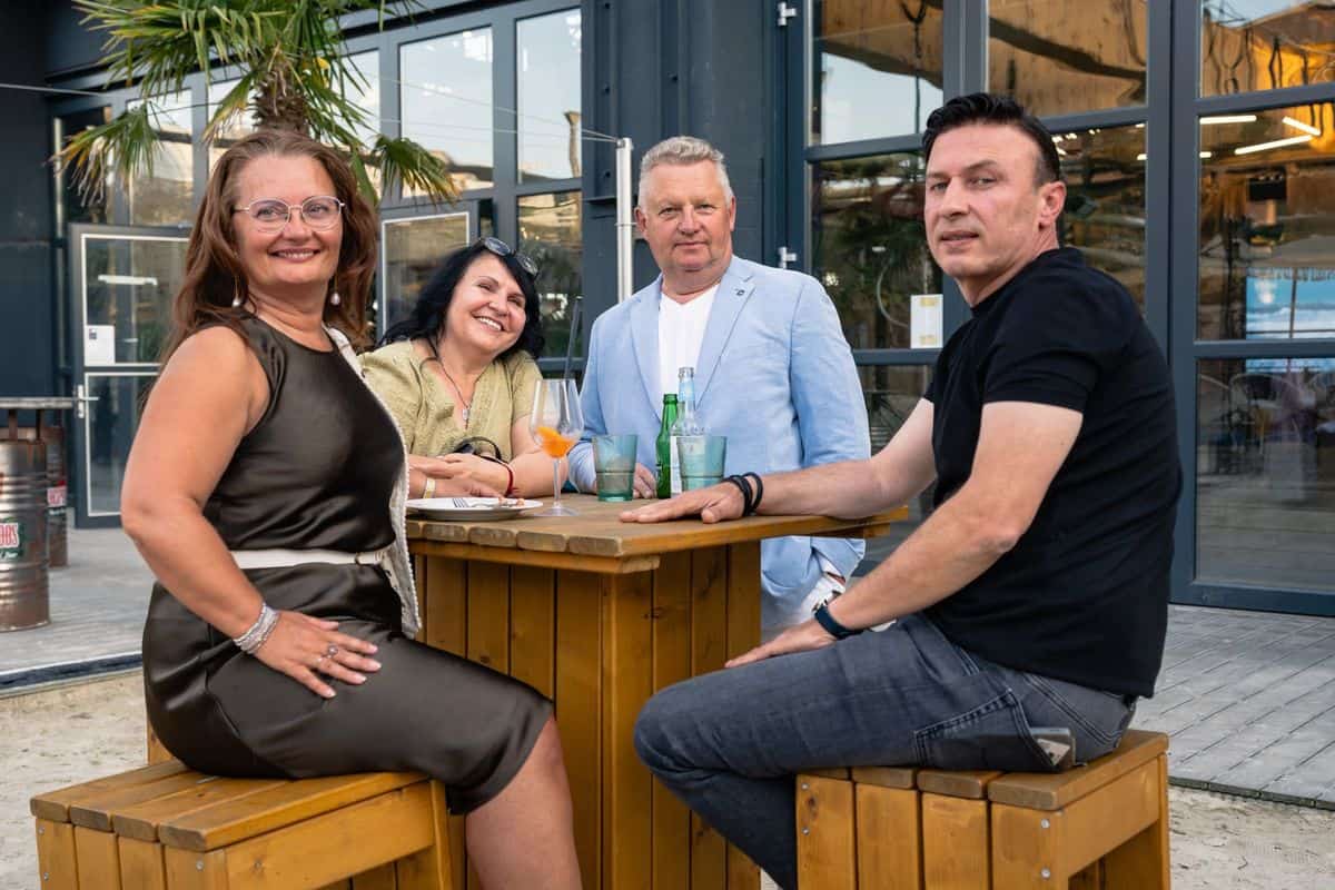 Vier Personen sitzen an einem Holztisch im Freien, umgeben von modernen Gebäuden und Palmen, mit Getränken in der Hand.