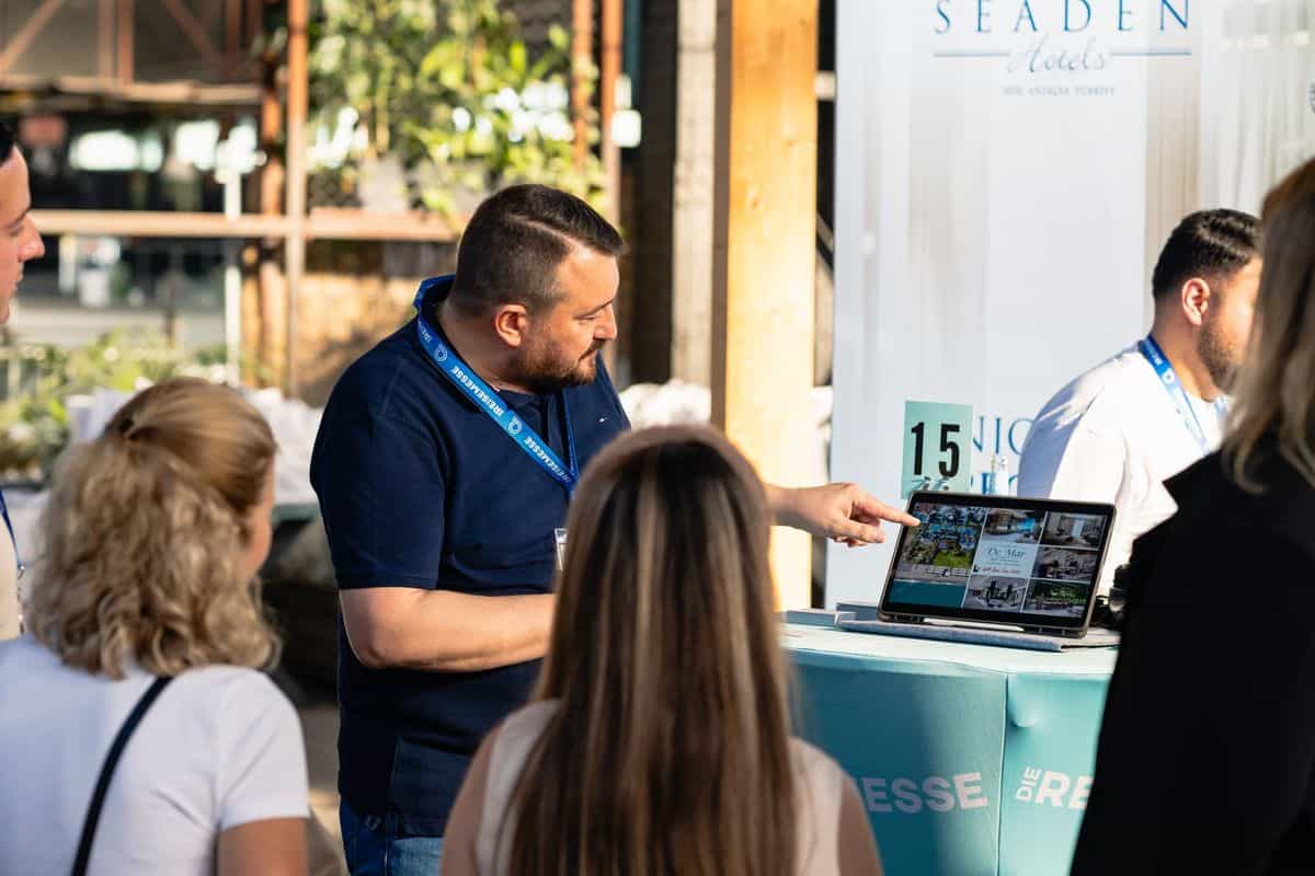 Ein Mann zeigt einer Gruppe von Personen auf einem Laptop Informationen, während sie an einem Messestand stehen, umgeben von Pflanzen.