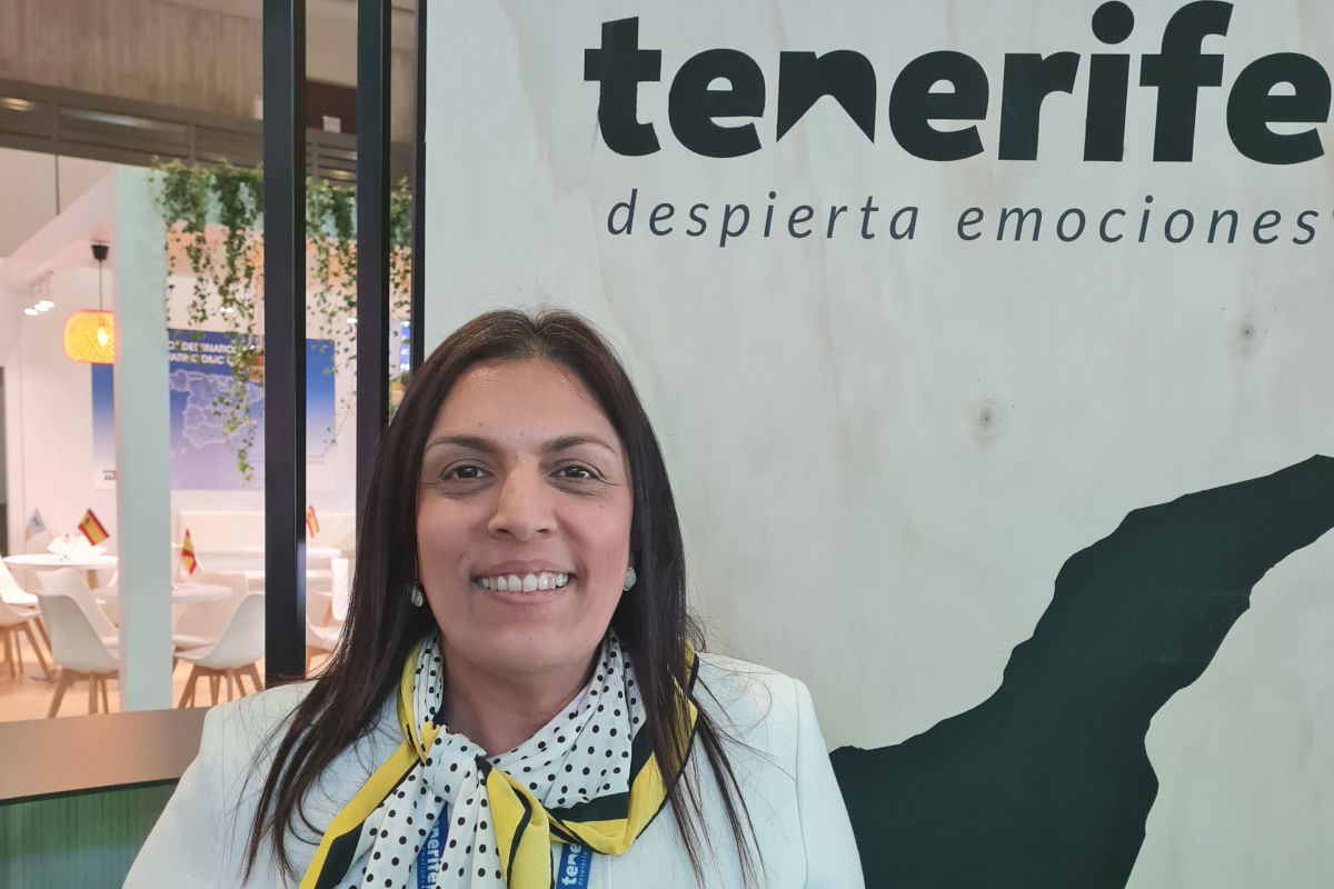 Eine lächelnde Frau mit dunklen, schulterlangen Haaren steht an einem Messestand vor einem großen Banner mit der Aufschrift „tenerife despierta emociones“ und einer Silhouette der Insel. Sie trägt ein helles Sakko, ein gepunktetes Halstuch in Gelb, Schwarz und Weiß sowie ein Namensband. Im Hintergrund sind Sitzgelegenheiten, Pflanzen und kleine Flaggen zu sehen.