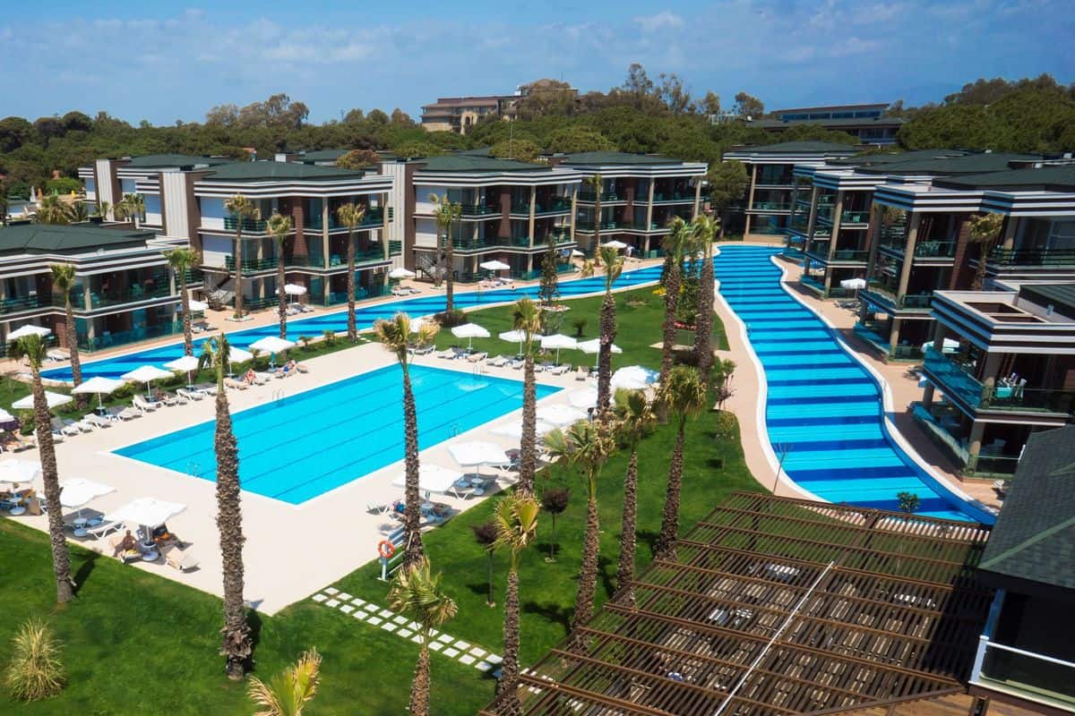 Luftaufnahme eines modernen Resorts mit mehreren Gebäuden, Palmen und einem großen Poolbereich mit Liegen und Sonnenschirmen.