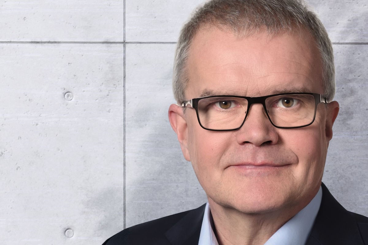Porträt eines älteren Mannes mit kurzen grauen Haaren und schwarzer Brille. Er trägt ein dunkles Sakko mit hellblauem Hemd und blickt neutral in die Kamera. Im Hintergrund ist eine graue Betonwand zu sehen.