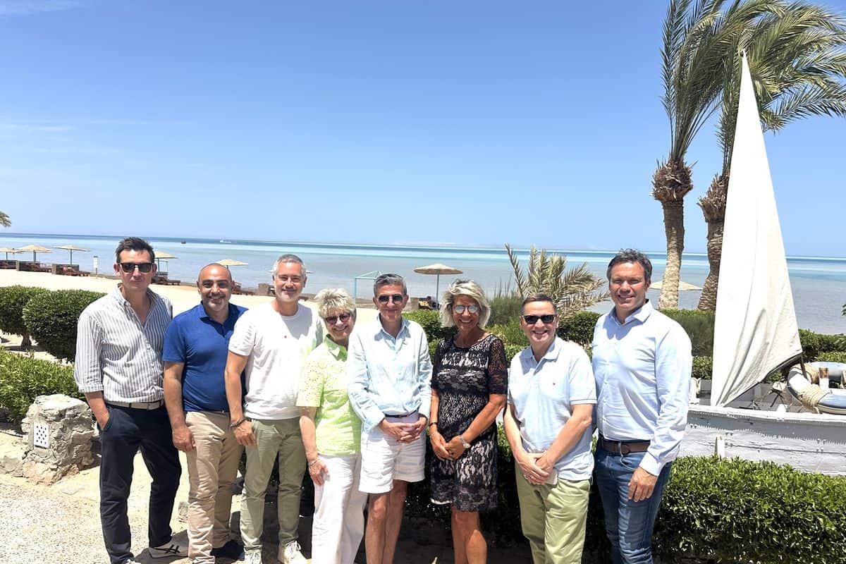 Eine Gruppe von acht Personen steht am Strand, umgeben von Palmen und Sonnenschirmen, mit Blick auf das Meer und den blauen Himmel.