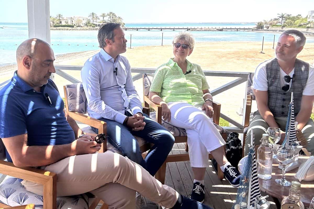 Vier Personen sitzen auf einer Terrasse mit Blick auf das Meer. Die Umgebung ist hell und einladend, mit Palmen im Hintergrund.