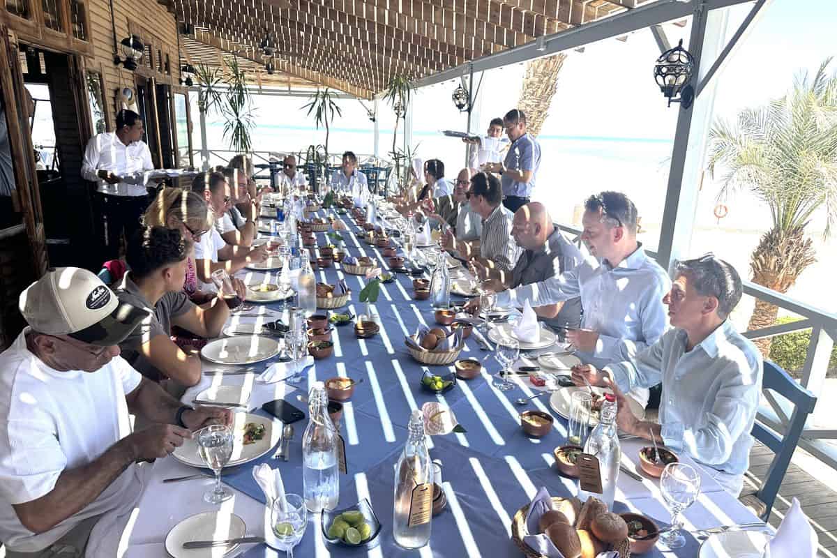 Eine lange Tafel mit Gästen, die an einem sonnigen Tag in einem Restaurant am Strand speisen. Wasserflaschen und Teller sind sichtbar.