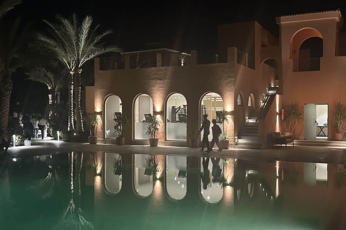 Ein modernes Gebäude mit beleuchteten Bögen und Palmen, reflektiert im Wasser eines Pools bei Nacht, umgeben von einer eleganten Atmosphäre.