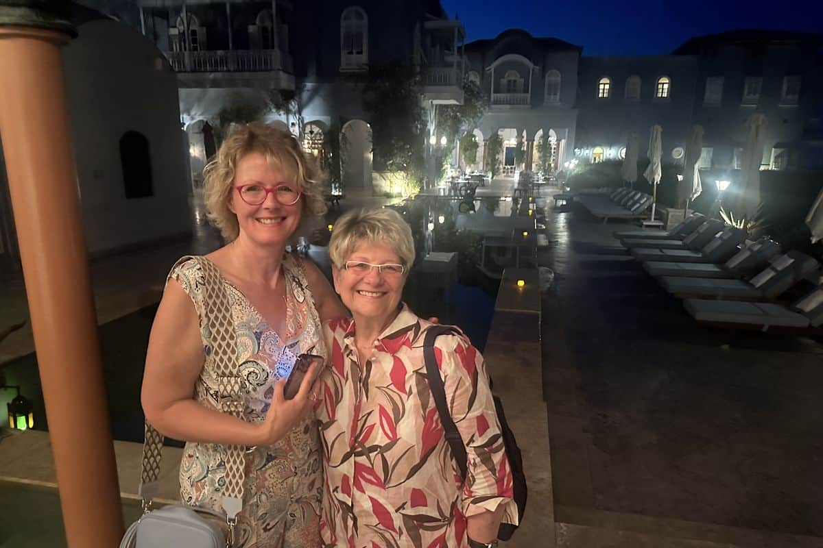 Zwei Frauen stehen lächelnd vor einem beleuchteten Hotelbereich mit einem Pool und Liegen im Hintergrund, bei Nacht aufgenommen.
