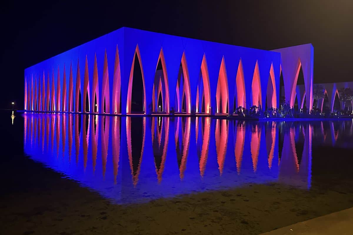 Moderne Architektur eines beleuchteten Gebäudes mit bunten Lichtern, reflektiert im Wasser bei Nacht.