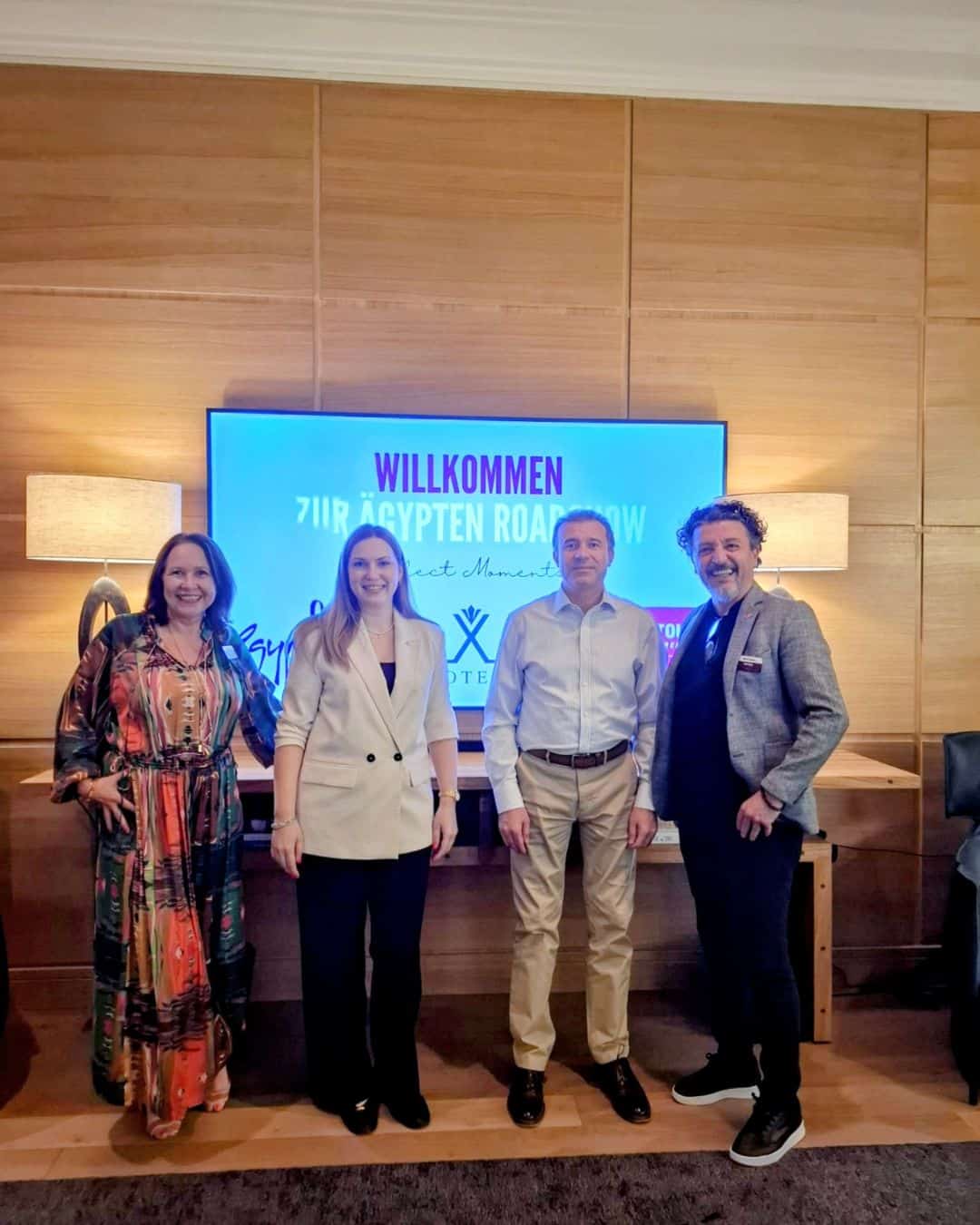 Vier Personen stehen vor einem Bildschirm mit der Aufschrift 'Willkommen zur Ägypten Roadshow' in einem modernen Innenraum.
