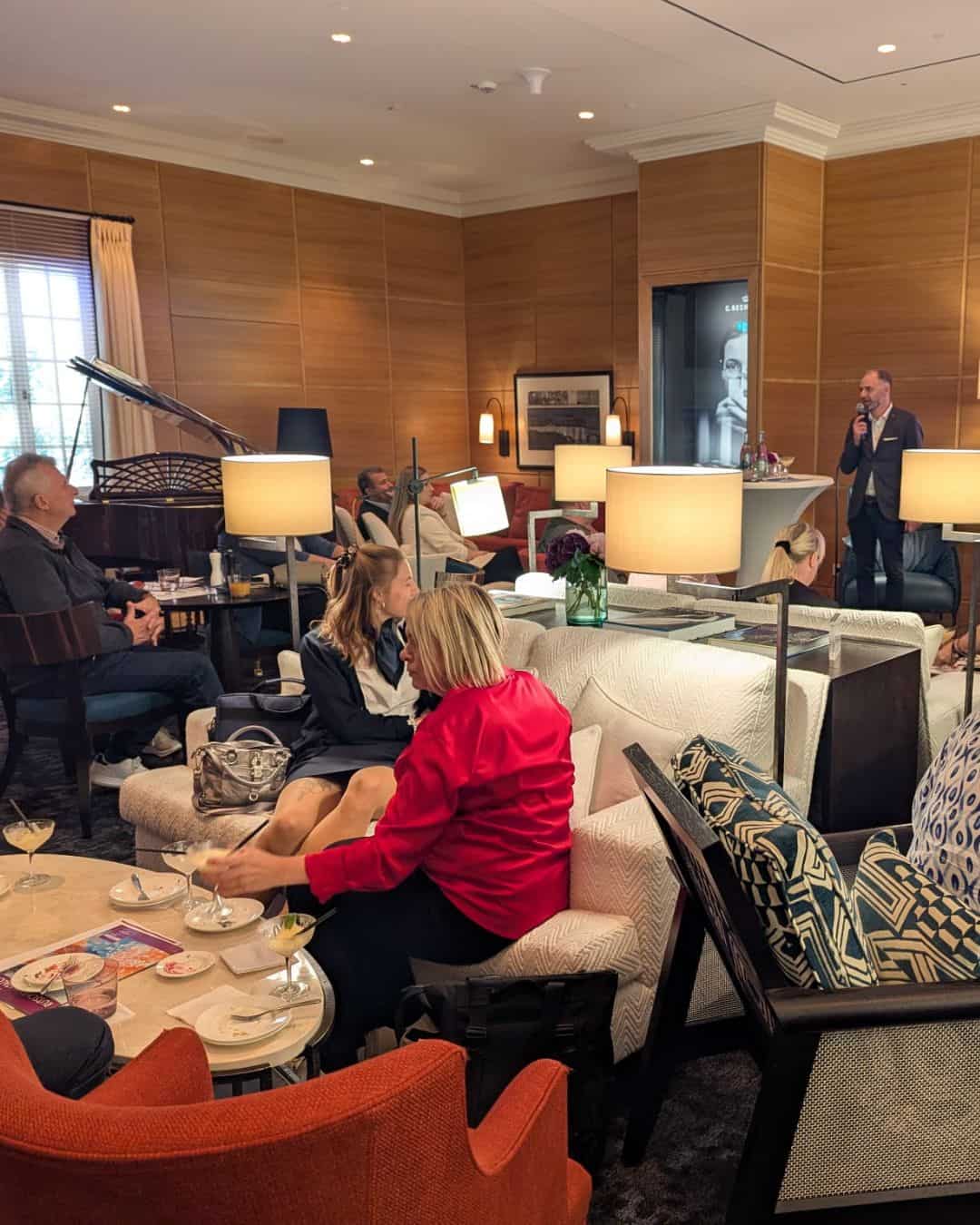 Eine elegante Lounge mit Gästen, die an Tischen sitzen. Eine Person spricht am Mikrofon, während andere entspannen und Getränke genießen.