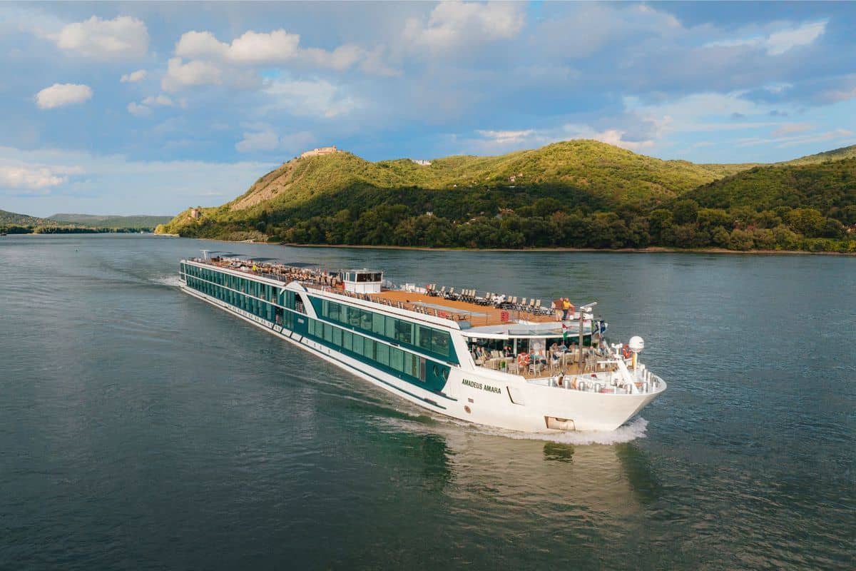 Ein modernes Flusskreuzfahrtschiff fährt auf einem ruhigen Gewässer, umgeben von grünen Hügeln und einem klaren Himmel mit wenigen Wolken.