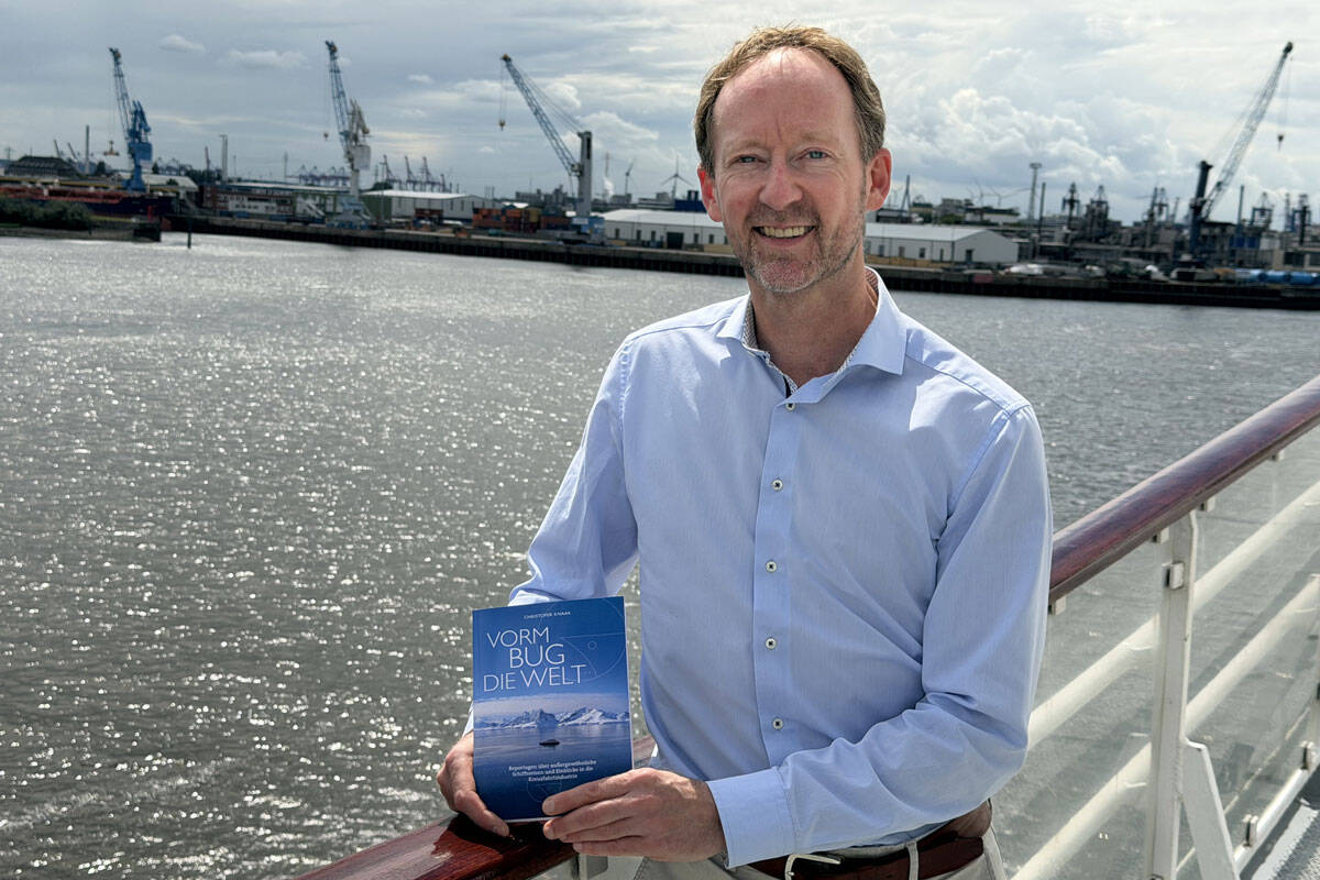 Ein Mann steht auf einem Schiff und hält ein Buch in der Hand, während im Hintergrund ein Hafen mit Kränen und Wasser sichtbar ist.