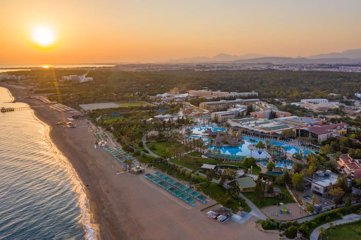 Luftaufnahme eines Resorts am Meer bei Sonnenuntergang, umgeben von Palmen und einem Poolbereich, mit einem langen Sandstrand im Vordergrund.