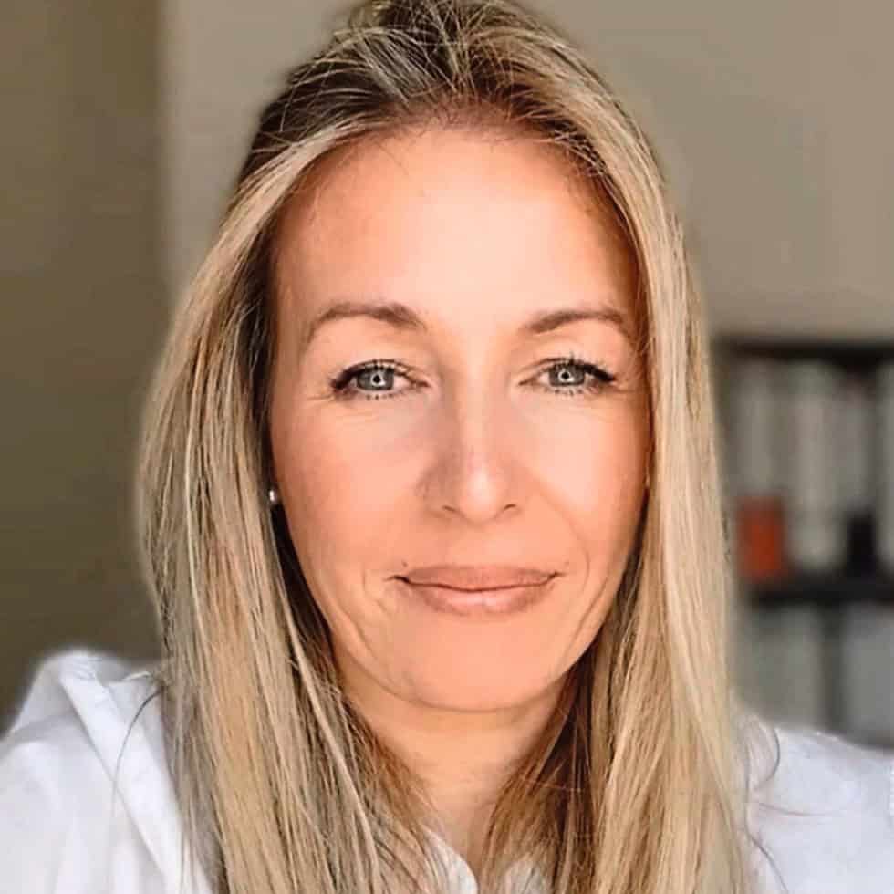 Porträt einer Frau mit langen blonden Haaren und einem freundlichen Lächeln, vor einem neutralen Hintergrund mit verschwommenen Regalen.