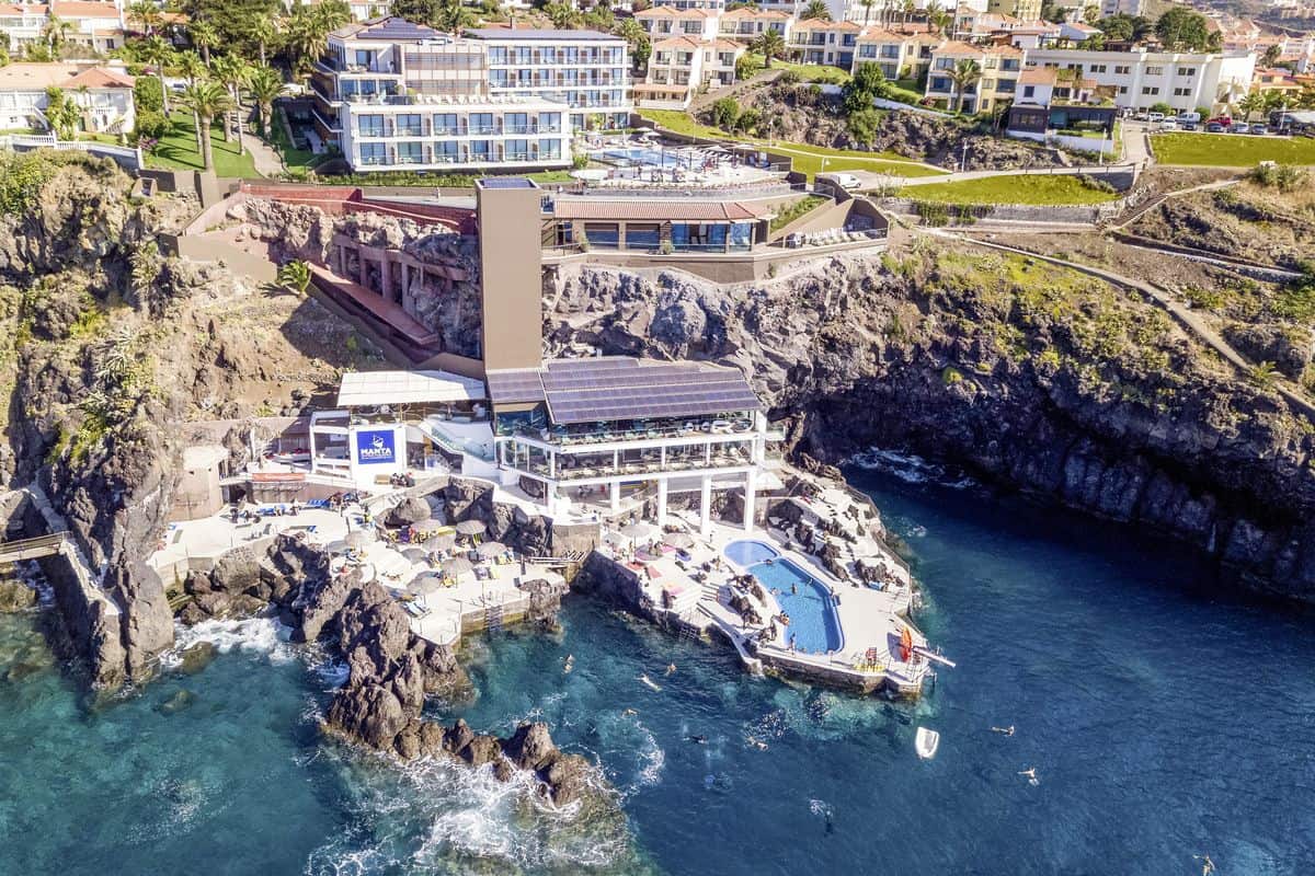 Luftaufnahme eines modernen Hotels an einer Küste mit einem Swimmingpool und Liegen auf den Felsen, umgeben von klarem Wasser.