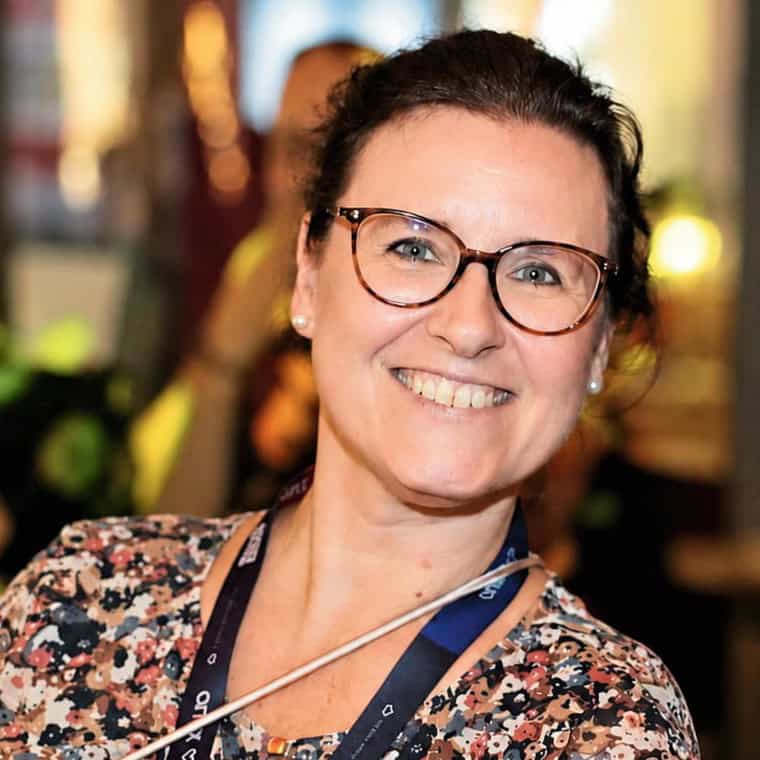 Eine lächelnde Frau mit Brille und Blumenmusterbluse, die in einem gut beleuchteten Raum steht, mit unscharfen Personen im Hintergrund.