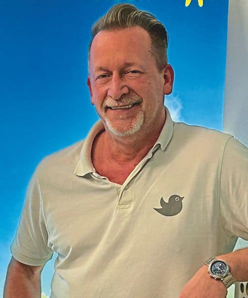 Ein Mann mit einem freundlichen Lächeln trägt ein helles Polohemd mit einem Vogelmotiv. Der Hintergrund zeigt einen klaren blauen Himmel.