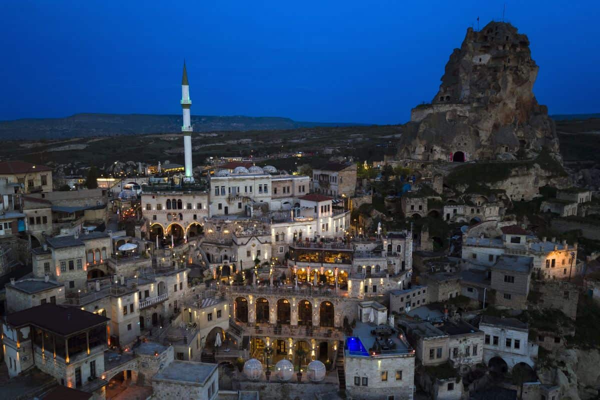 Luftaufnahme einer historischen Stadt mit beleuchteten Gebäuden, einer Moschee und einer Felsformation im Hintergrund bei Dämmerungslicht.